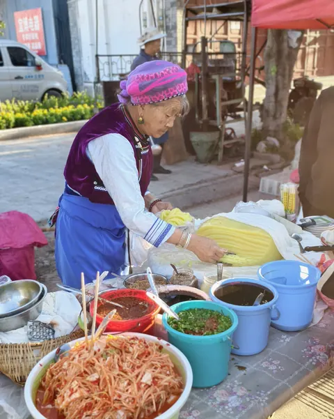 A Bai grandmother in traditional headwear selling homemade snacks at a village market